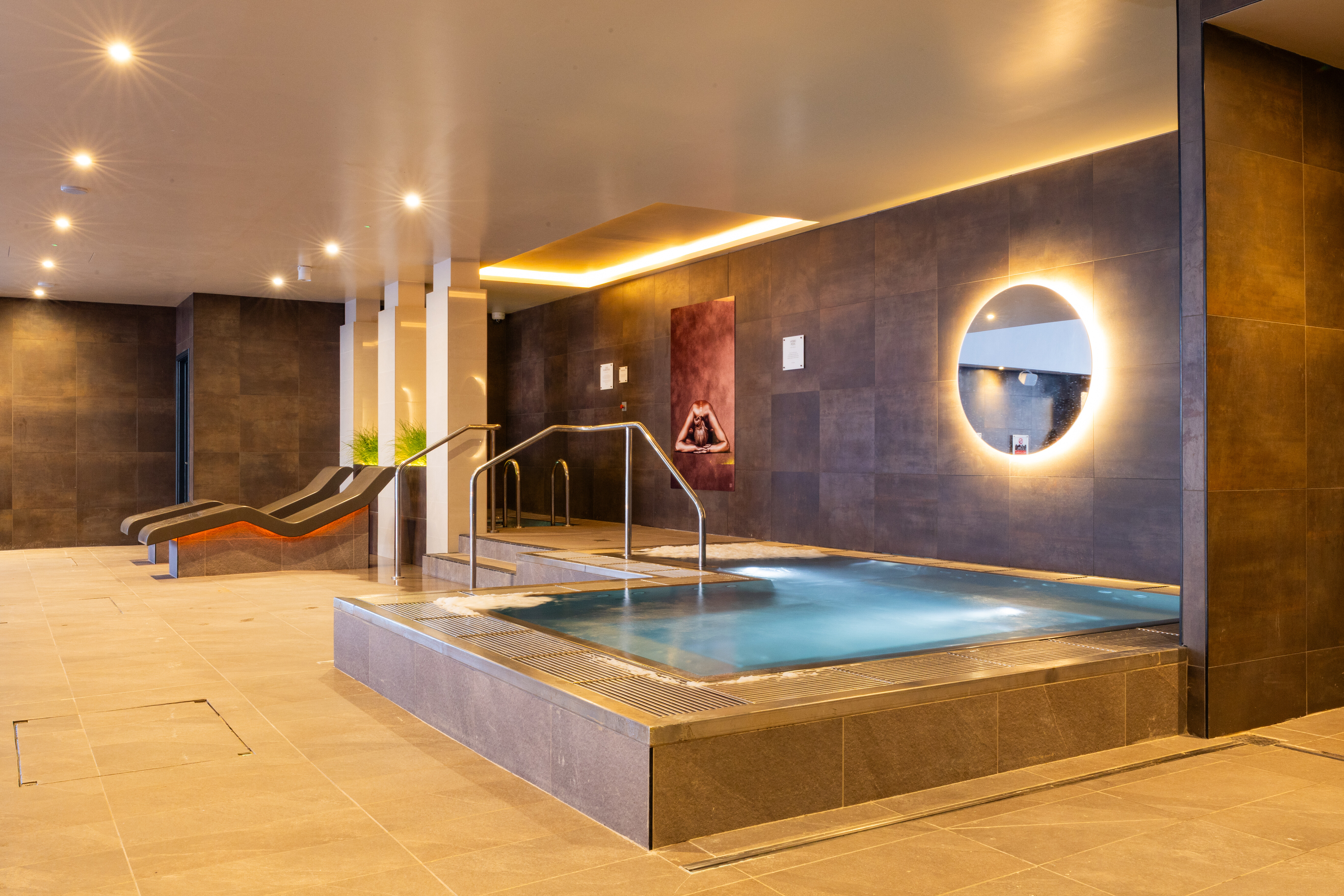 A jacuzzi underneath a backlit mirror with heated loungers in the background