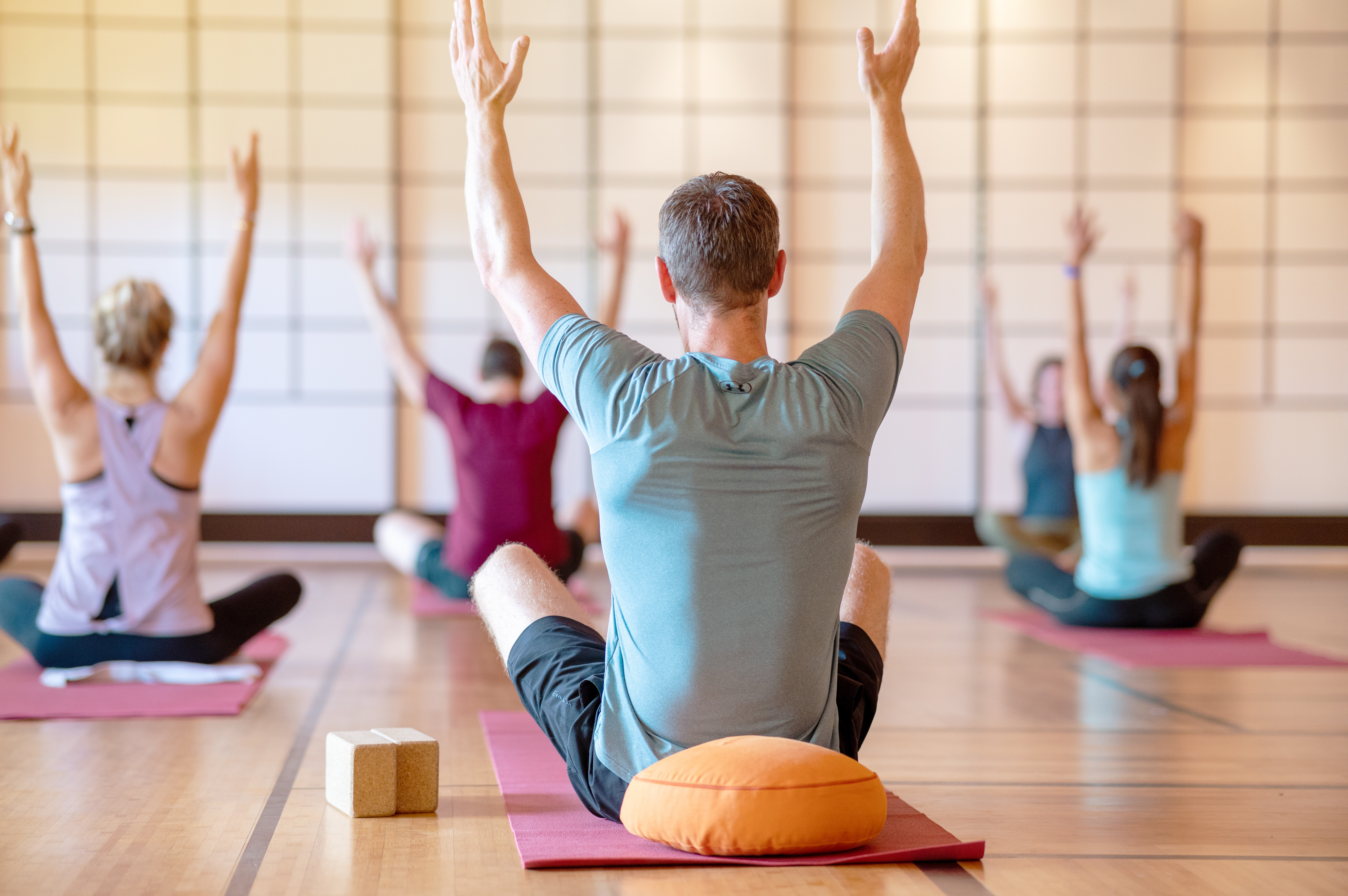 A group sits on yoga mats with arms stretched up, seen from behind