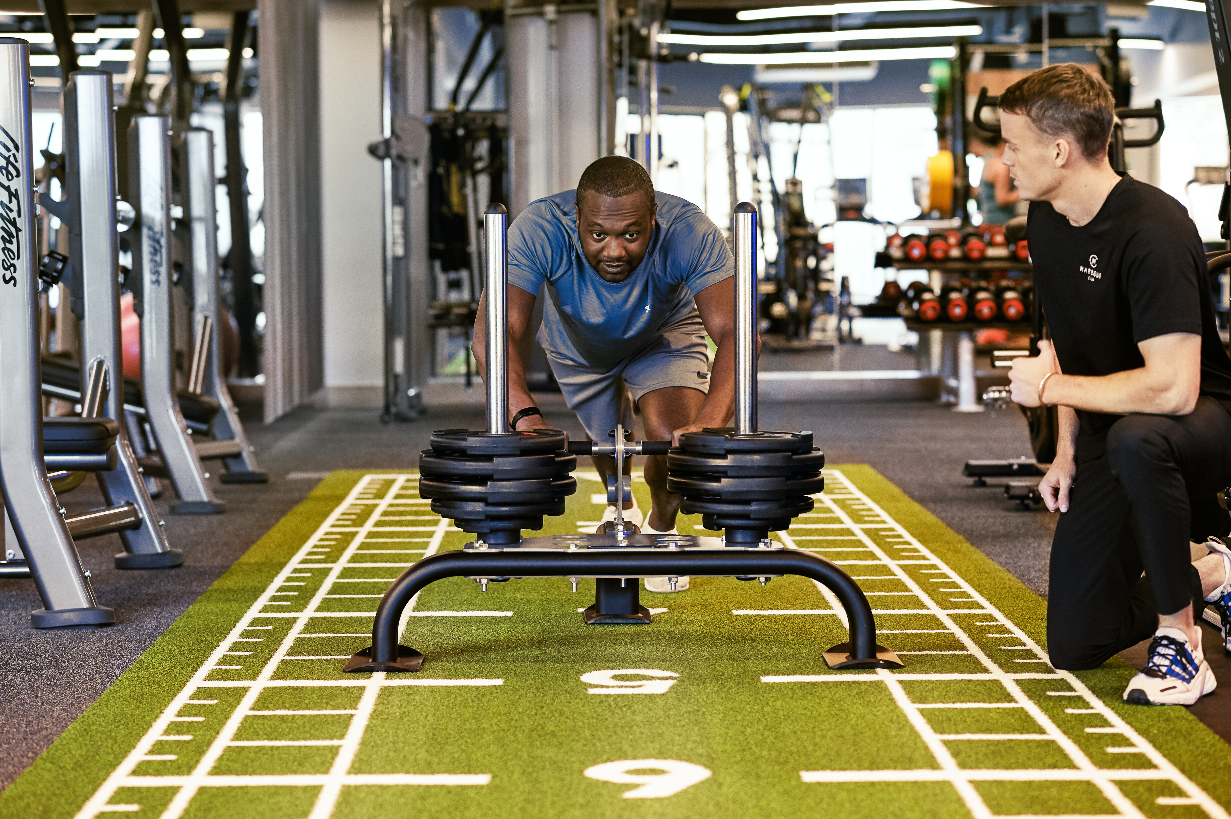 A man using a push sled with his personal trainer encouraging him