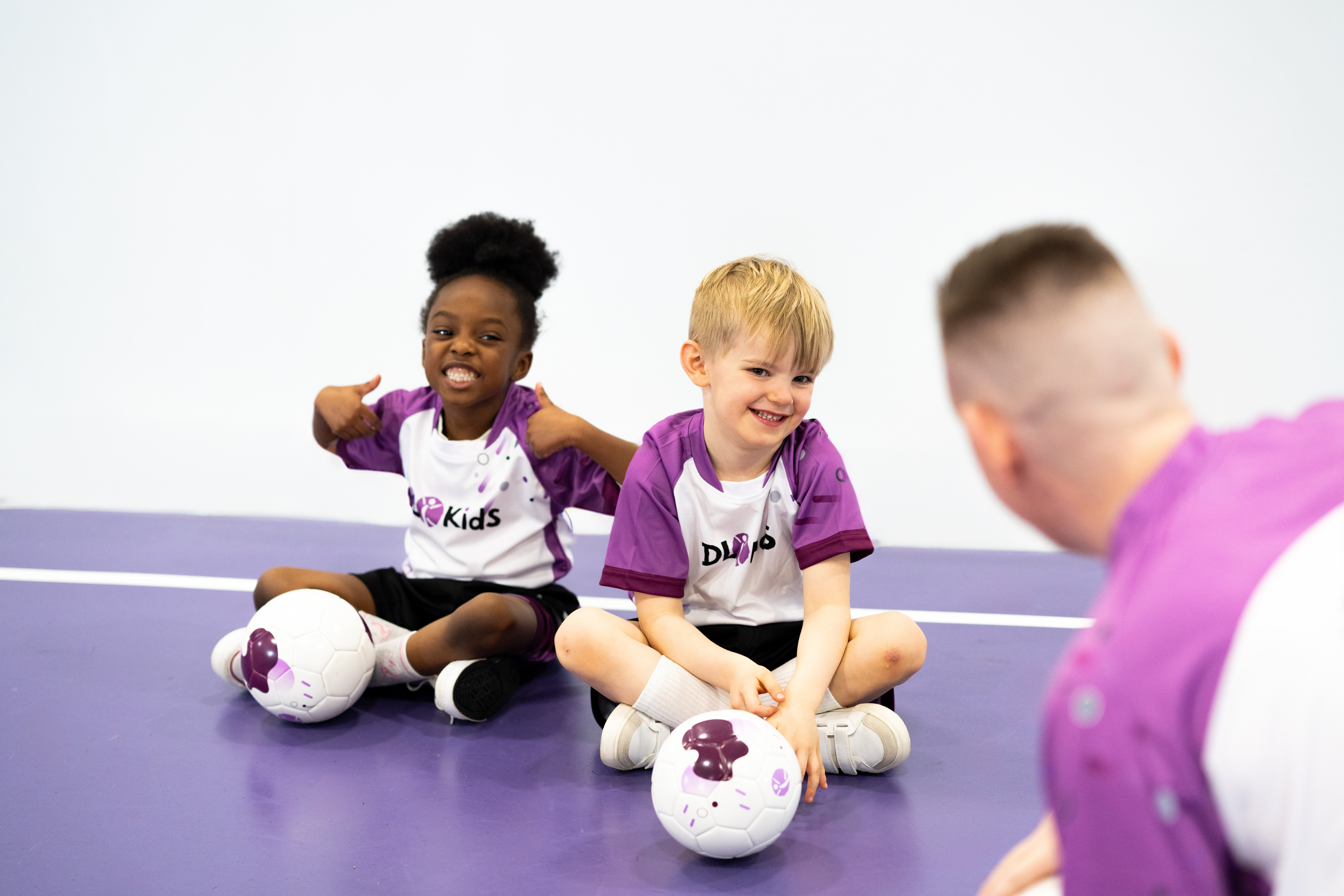 Children wearing DL Kids T-shirts sit on the floor smiling as they listen to their instructor