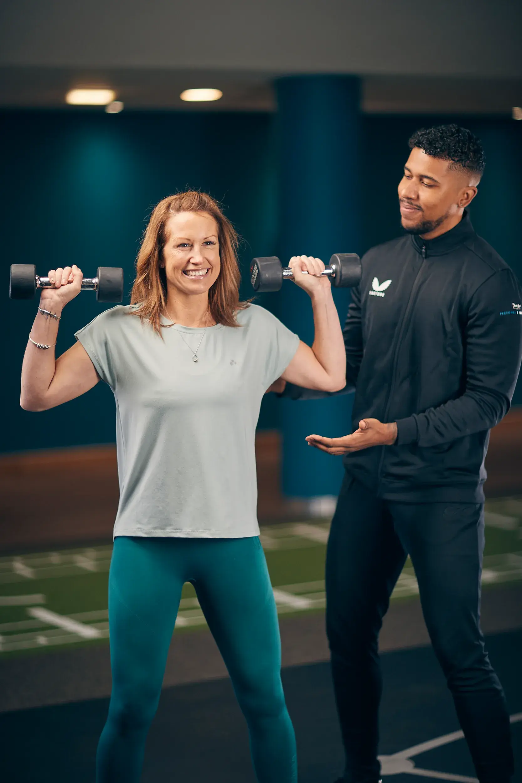 A woman lifts weights above her shoulders as her PT encourages her