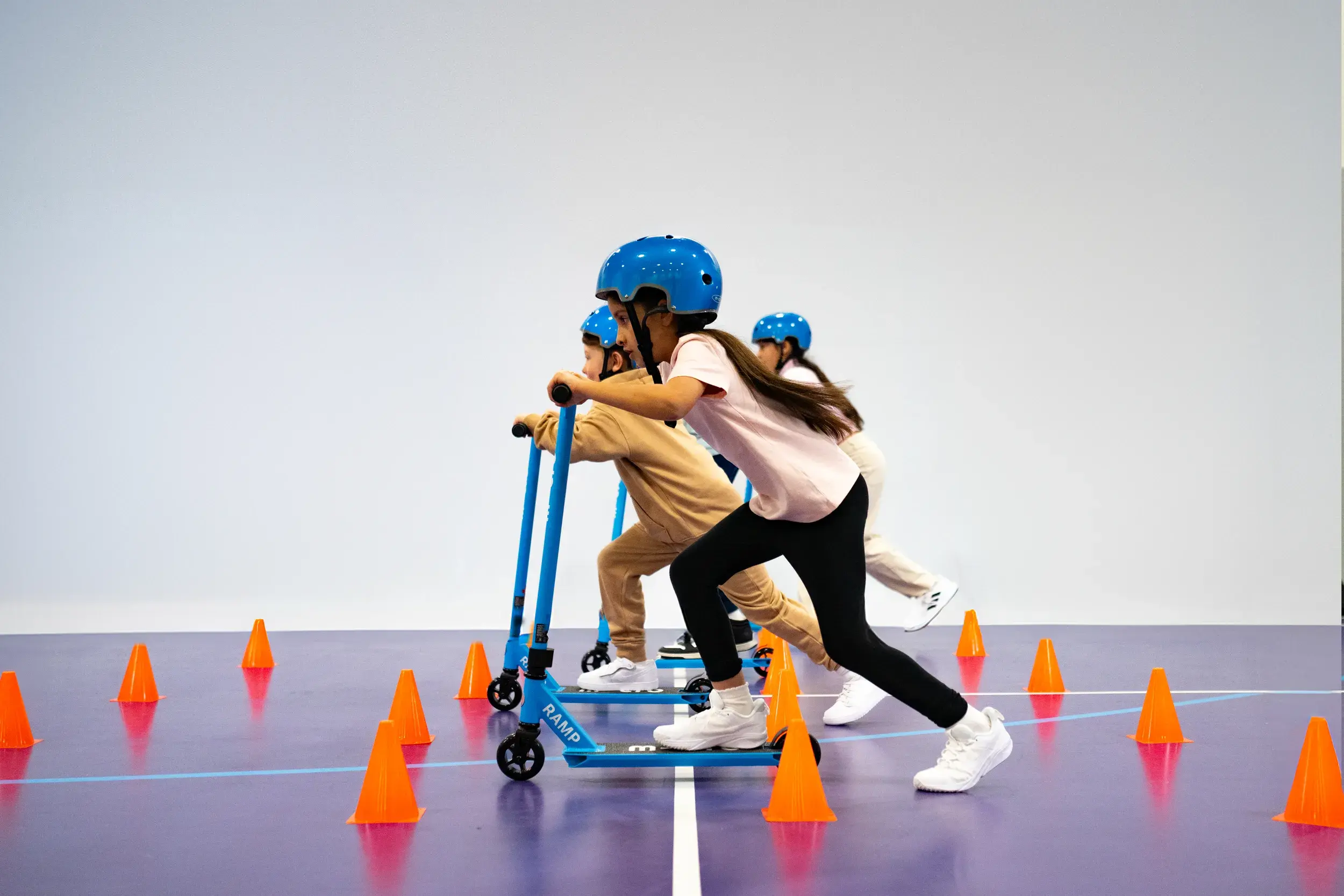 Children on scooters scoot between cones while wearing helmets.